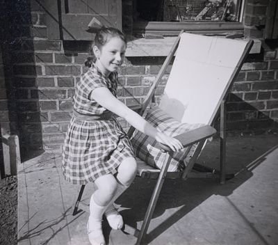 A girl sits on the edge of a wooden deckchair and smiles.