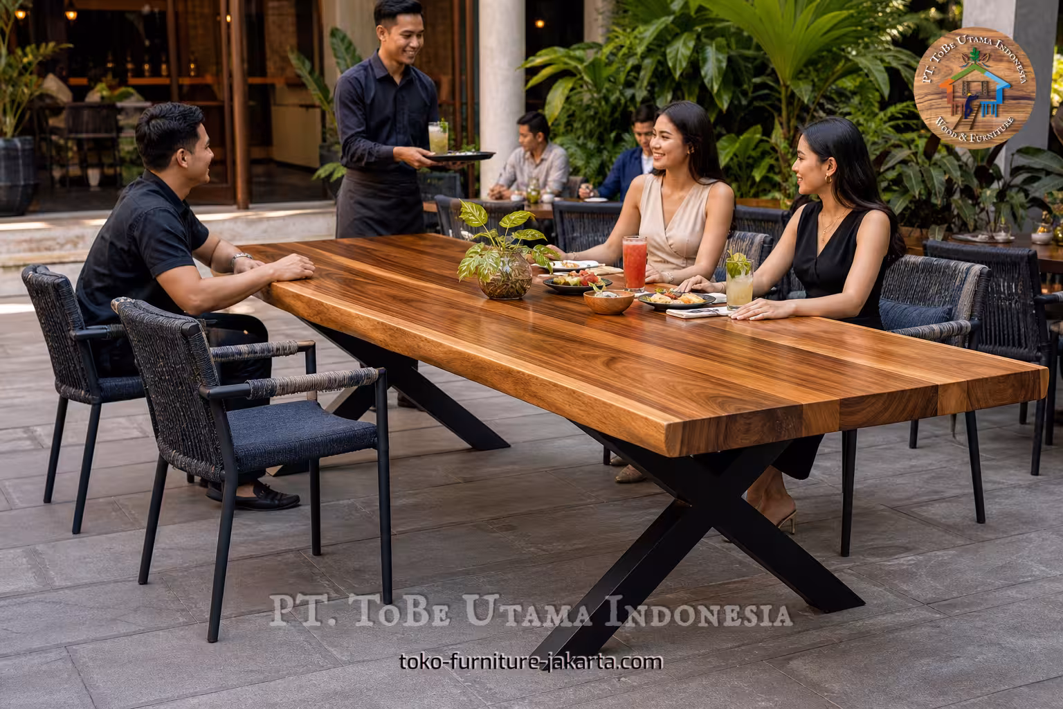 Meja makan kayu besar di kafe luar ruangan dengan para tamu, minuman, piring, dan tanaman yang rimbun.
