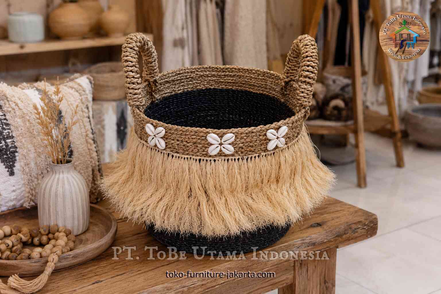 Round woven basket with black base, loop handles, raffia fringe, and white shell accents on a wooden table